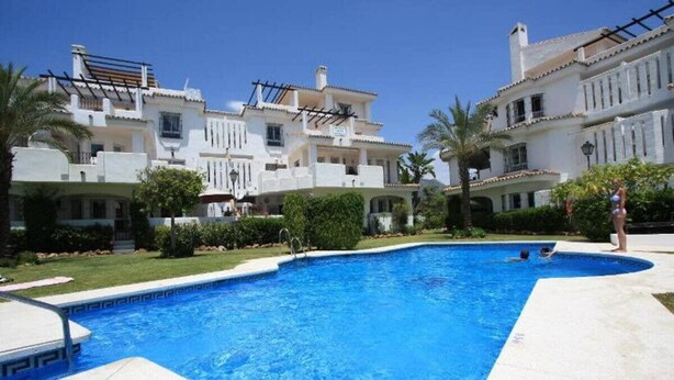 Una piscina rodeada de edificios blancos con balcones, un cielo azul despejado y un césped bien cuidado.