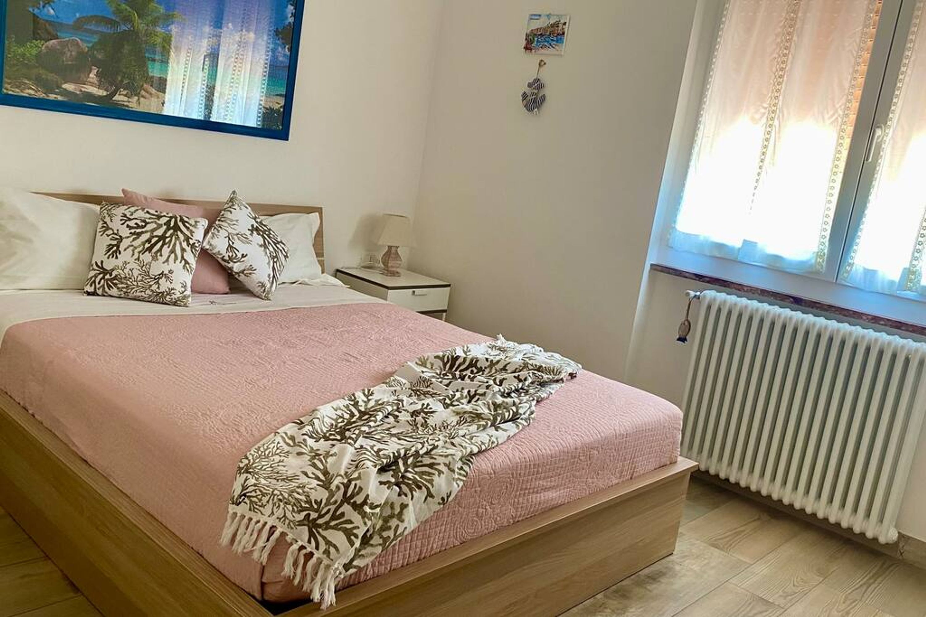 A bedroom with a bed, pillows, a nightstand, a lamp, a window with curtains, and a radiator.
