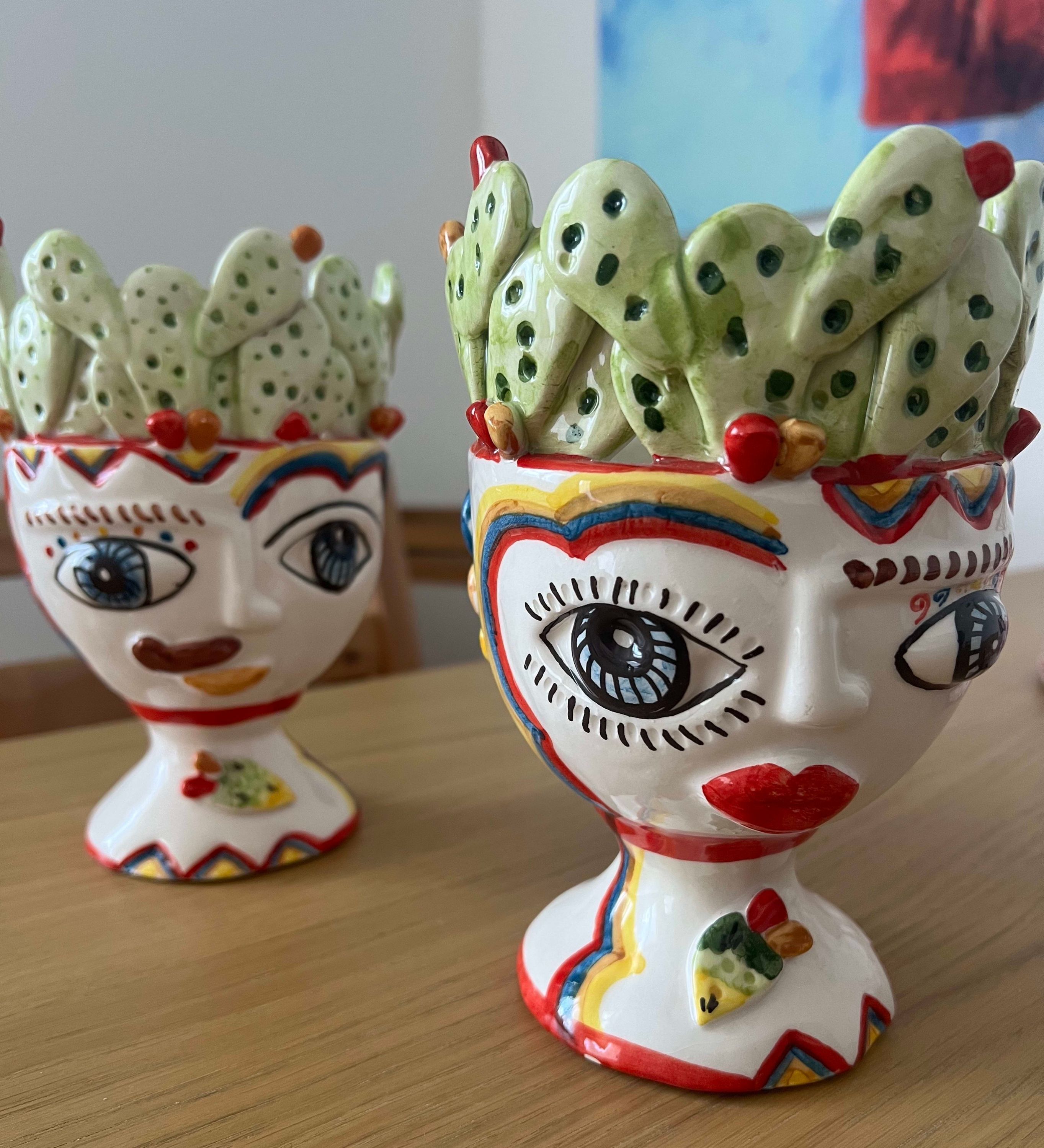Deux vases en céramique ornés de visages, l’un avec un œil bleu et l’autre avec un œil rouge, sont posés sur une surface en bois.