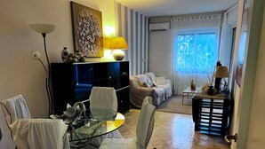 A modern living room with a glass dining table, white chairs, a sofa, and a coffee table. There is a painting on the wall, a lamp, and a window with curtains. - Rebecca s Sanremo Collection I (Sanremo)