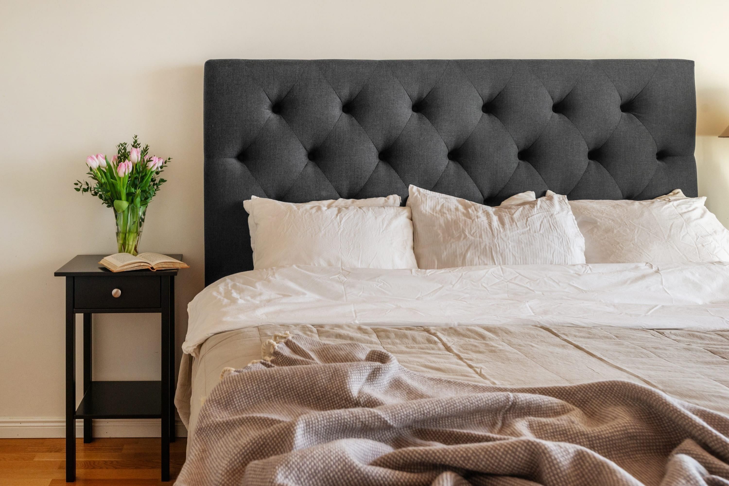 Una cama con cabecera capitonada, ropa de cama blanca, una mesita de noche con un libro y flores, y una lámpara.