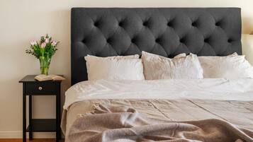 Una cama con cabecera capitonada, ropa de cama blanca, una mesita de noche con un libro y flores, y una lámpara.