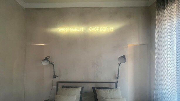 Un dormitorio con una cama, dos lámparas de noche y un letrero de neón que indica 'WEST BERLIN EAST BERLIN'.