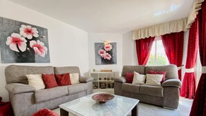 A living room with two grey sofas, red cushions, a glass coffee table, and floral paintings on the walls.