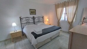 A neatly made bed with white linens and a dark blanket, a bedside lamp, a small wooden nightstand, and a framed picture on the wall.