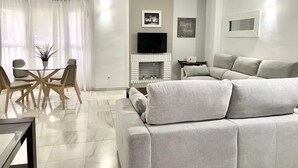 A modern living room with a grey sofa, a glass dining table with chairs, a fireplace, and a flat-screen TV mounted on the wall.