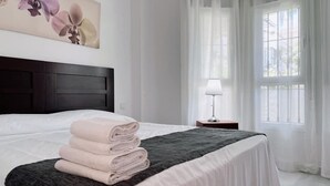 A neatly made bed with folded towels, a dark headboard, a bedside table with a lamp, and a window with sheer curtains. - Spacious Poolside 3 Bedroom F-5 (Marbella)