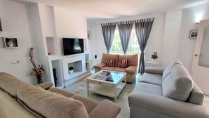 A modern living room with a beige sofa set, a glass coffee table, a flat-screen TV mounted on a white wall, and a fireplace. - Villa Emma 4 Bedrooms F-3 (Marbella)