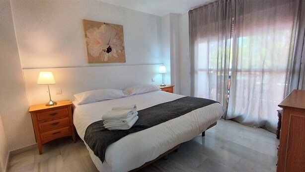 A hotel room with a large bed, two bedside lamps, a nightstand, a wooden dresser, and a window with sheer curtains. - Villa Emma 4 Bedrooms F-3 (Marbella)