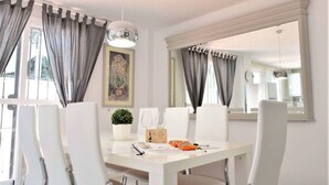 A dining area with a white table, four chairs, a potted plant, and a painting on the wall. - Villa Emma 4 Bedrooms F-3 (Marbella)