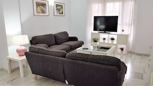 Una sala de estar moderna con un sofá seccional gris, una mesa de centro blanca, un televisor y una pequeña mesita auxiliar blanca con una lámpara rosa.