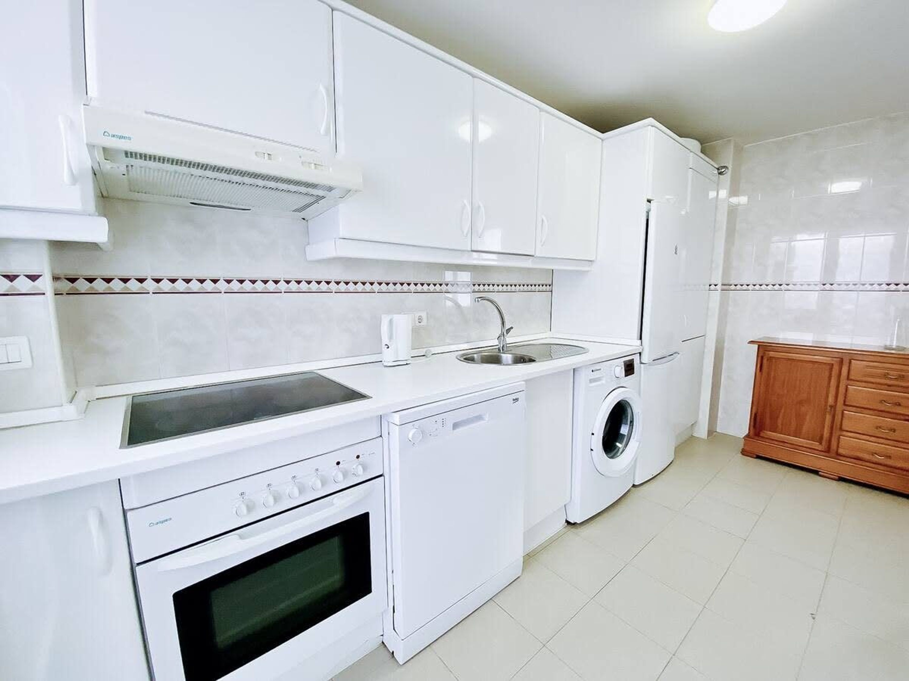 Une cuisine moderne avec des meubles blancs, un four encastré, un lave-vaisselle, un lave-linge et un évier.