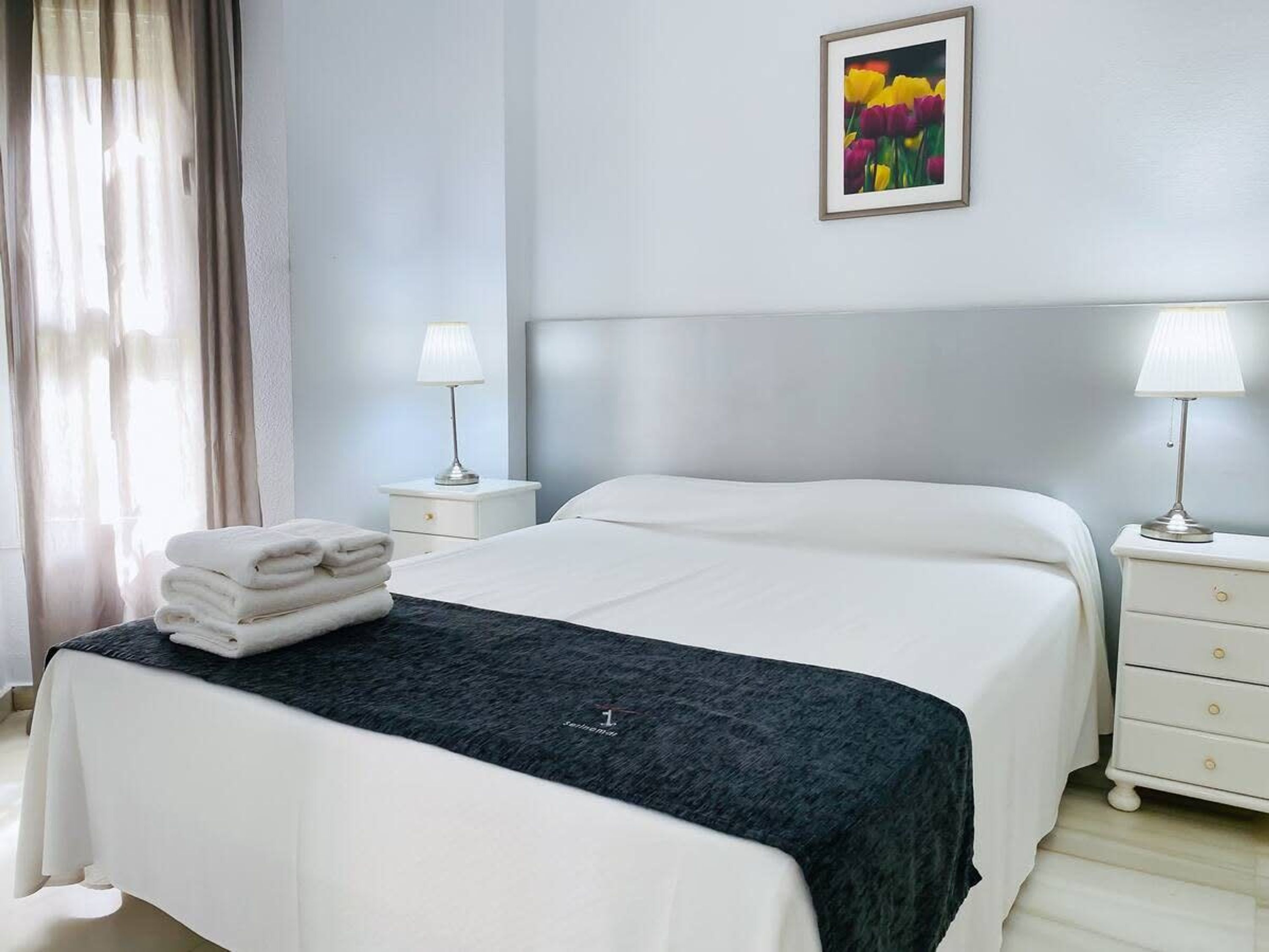 A neatly made bed with white linens and a dark blanket, two bedside tables with lamps, a framed picture on the wall, and a window with curtains.
