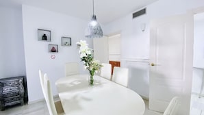 Un comedor con una mesa redonda, sillas blancas, una araña y un florero.