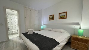A bedroom with a bed, a bedside table with a lamp, a wardrobe, and a door leading to a bathroom. - Standard 2 Bedroom Ground Floor Apartment F-3 (Marbella)