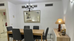 A dining area with a wooden table and six chairs, a chandelier, a mirror, and a side table with a lamp. - Standard 2 Bedroom Ground Floor Apartment F-3 (Marbella)