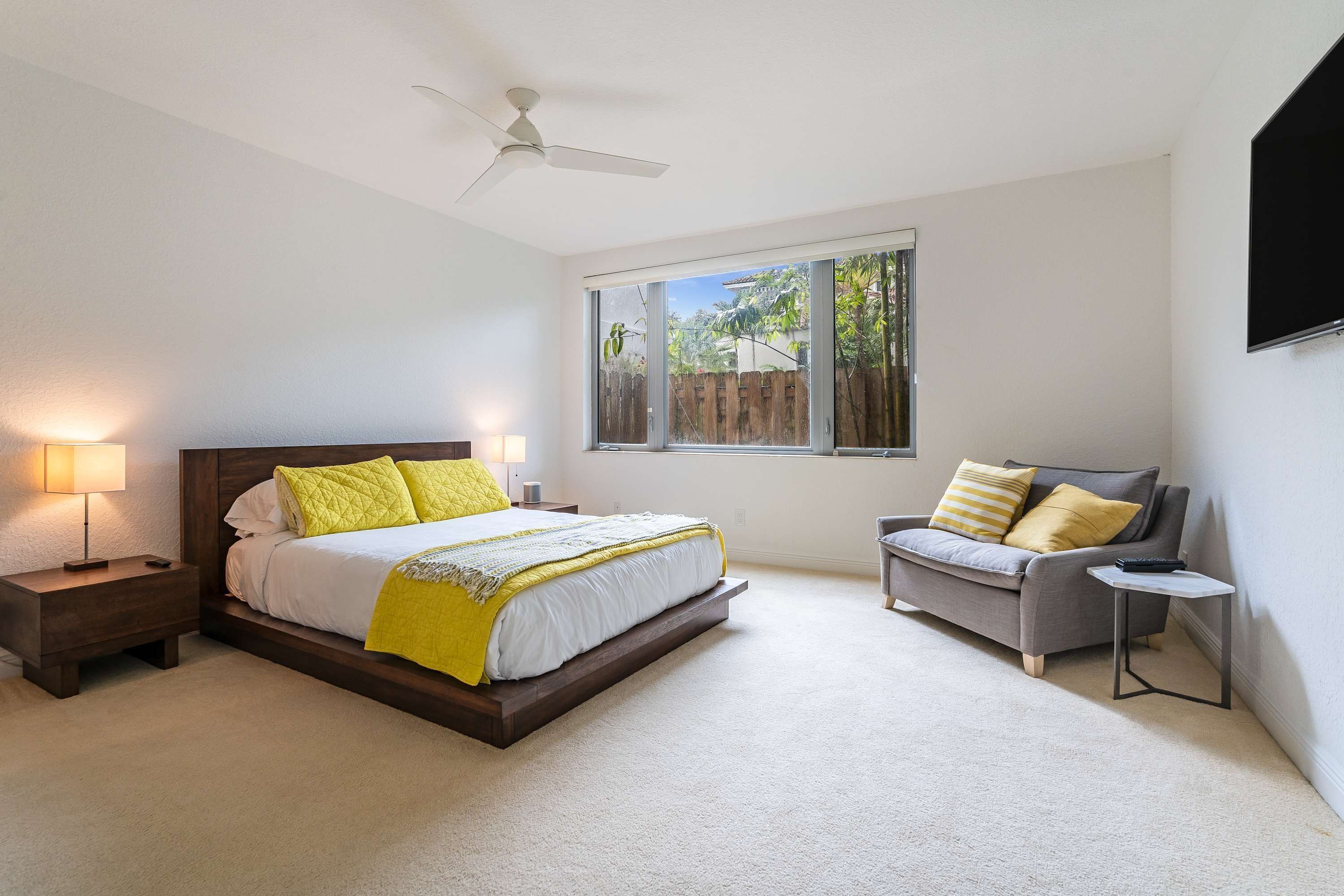 A bedroom with a bed, a nightstand, a chair, a small table, a ceiling fan, and a window with a view of greenery.