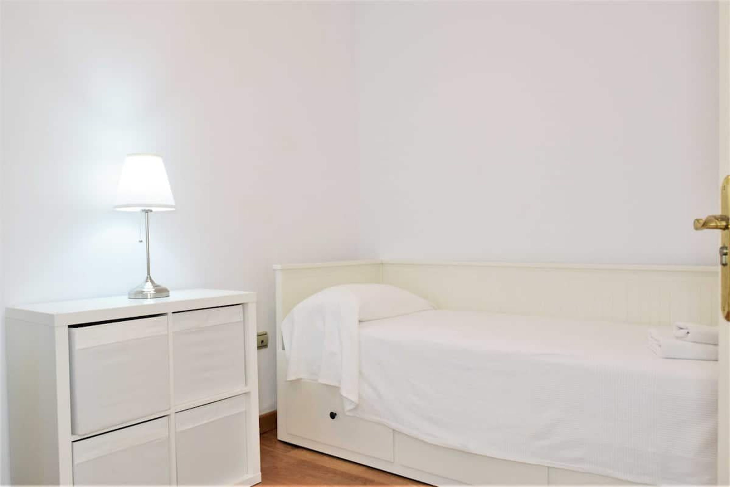 Una cama blanca con cabecera, una mesita de noche con lámpara y un armario blanco.