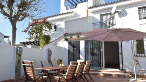 Un patio con una mesa y sillas de madera, un paraguas rosa y un edificio blanco con balcones.