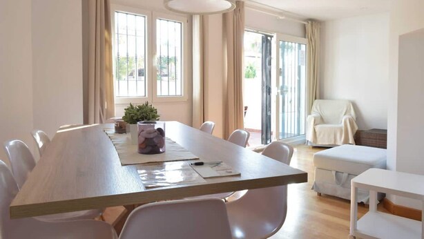 Un comedor moderno con una larga mesa, sillas blancas y una araña. Hay ventanas grandes con cortinas, un sillón blanco y una mesita con una planta.