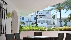 Un patio con mobiliario de mimbre, una mesa de vidrio y vistas a edificios blancos con balcones y palmeras.