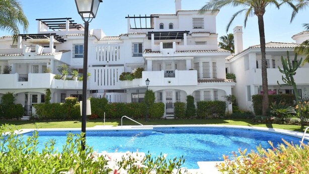 Un área de piscina con una piscina azul cristalina, rodeada de exuberante vegetación y edificios blancos con balcones.