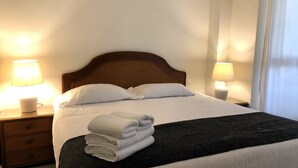 Una cama bien hecha con sábanas blancas y una pila de toallas dobladas, dos lámparas de noche y una mesita de noche de madera con un cajón.