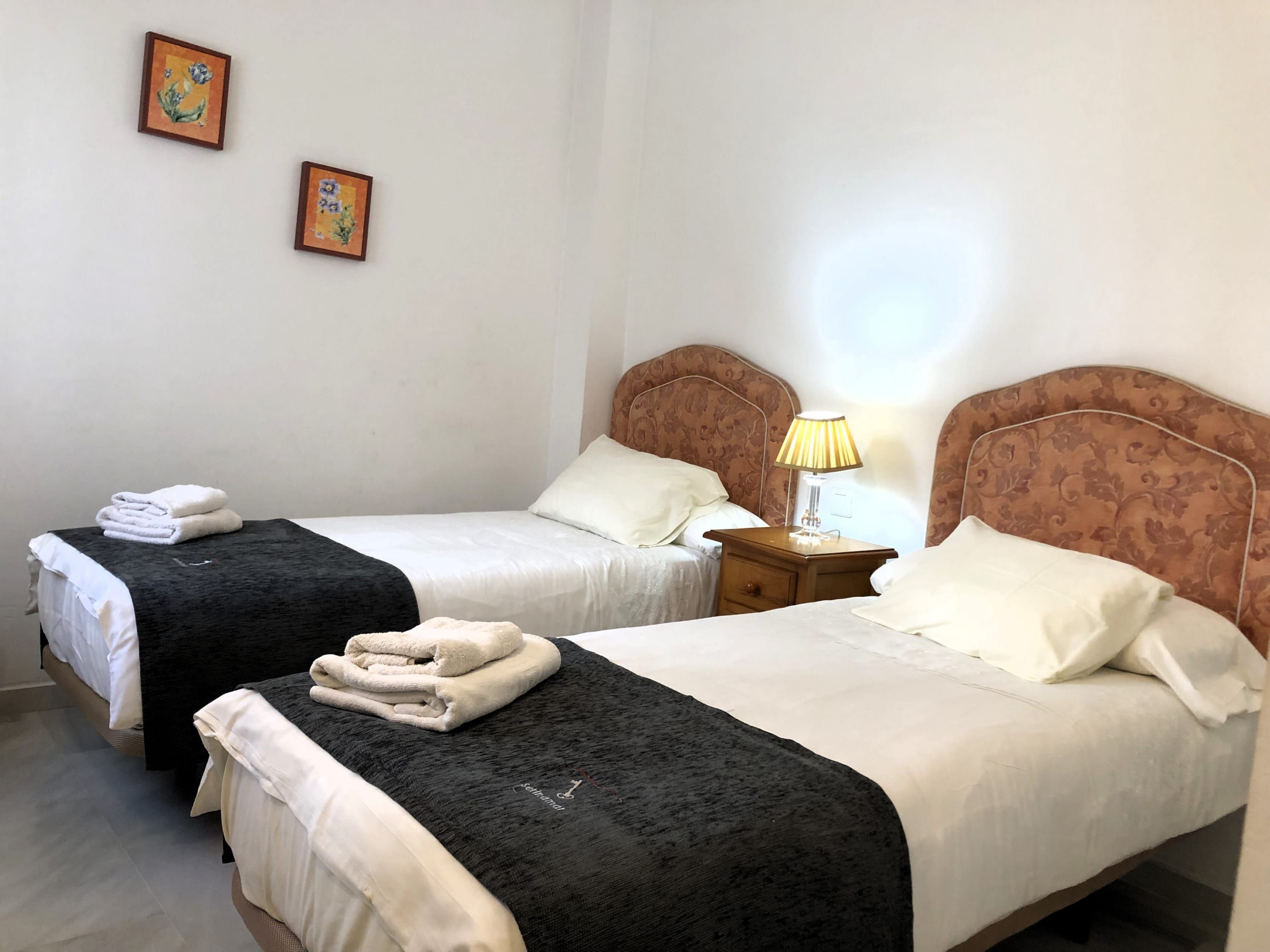 A hotel room with two beds, each with a black and white bedspread, white pillows, and folded towels. There is a nightstand with a lamp between the beds.