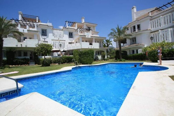 A swimming pool surrounded by white buildings with balconies and a clear blue sky. - Bright 2 Bedroom First Floor F-4 (Marbella)