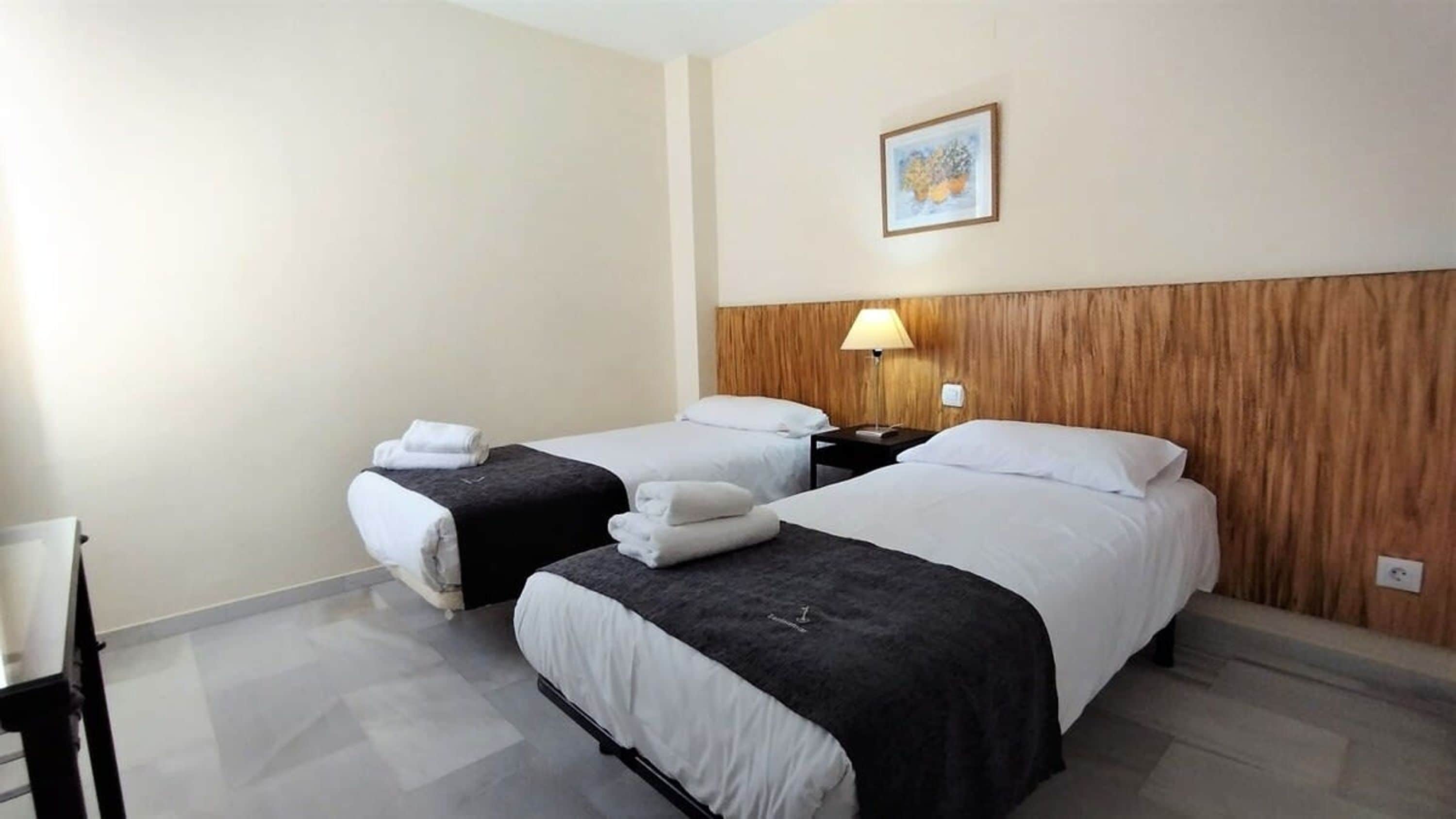 A hotel room with two beds, a wooden headboard, a nightstand with a lamp, and a framed picture on the wall.