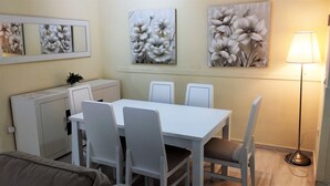 Un comedor con una mesa y sillas blancas, un mueble pequeño y tres cuadros con motivos florales en la pared.