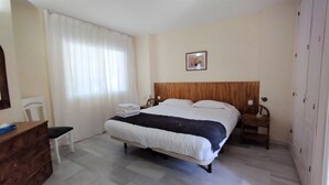 Una habitación de hotel con cama, cabecera de madera, mesita de noche, escritorio, silla y ventana con cortinas.