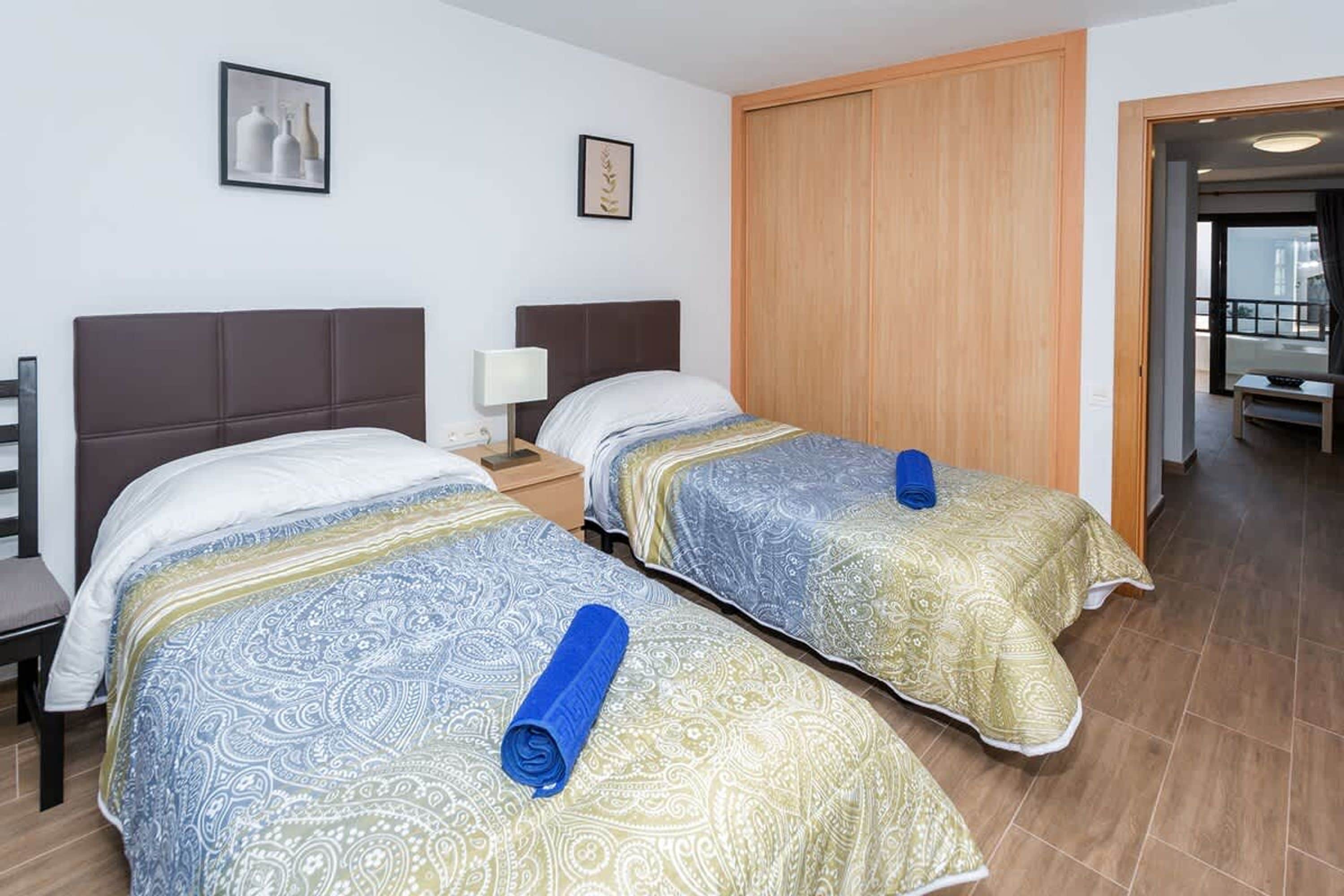 A hotel room with two beds, each with patterned bedding and a rolled towel. There is a nightstand with a lamp, a wooden wardrobe, and a view of the outside balcony.