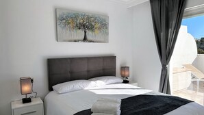 A bedroom with a large bed, a nightstand with a lamp, a painting of a tree, and a view of the outdoors. - Exceptional Views - Dama de Noche 1 Bedroom Penthouse (Marbella)