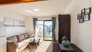 A modern living room with a sofa, coffee table, and a dining table. There is a sliding glass door leading to a balcony with a view of buildings. - Apartamento Golden Teguise 2 (Alcorcon)