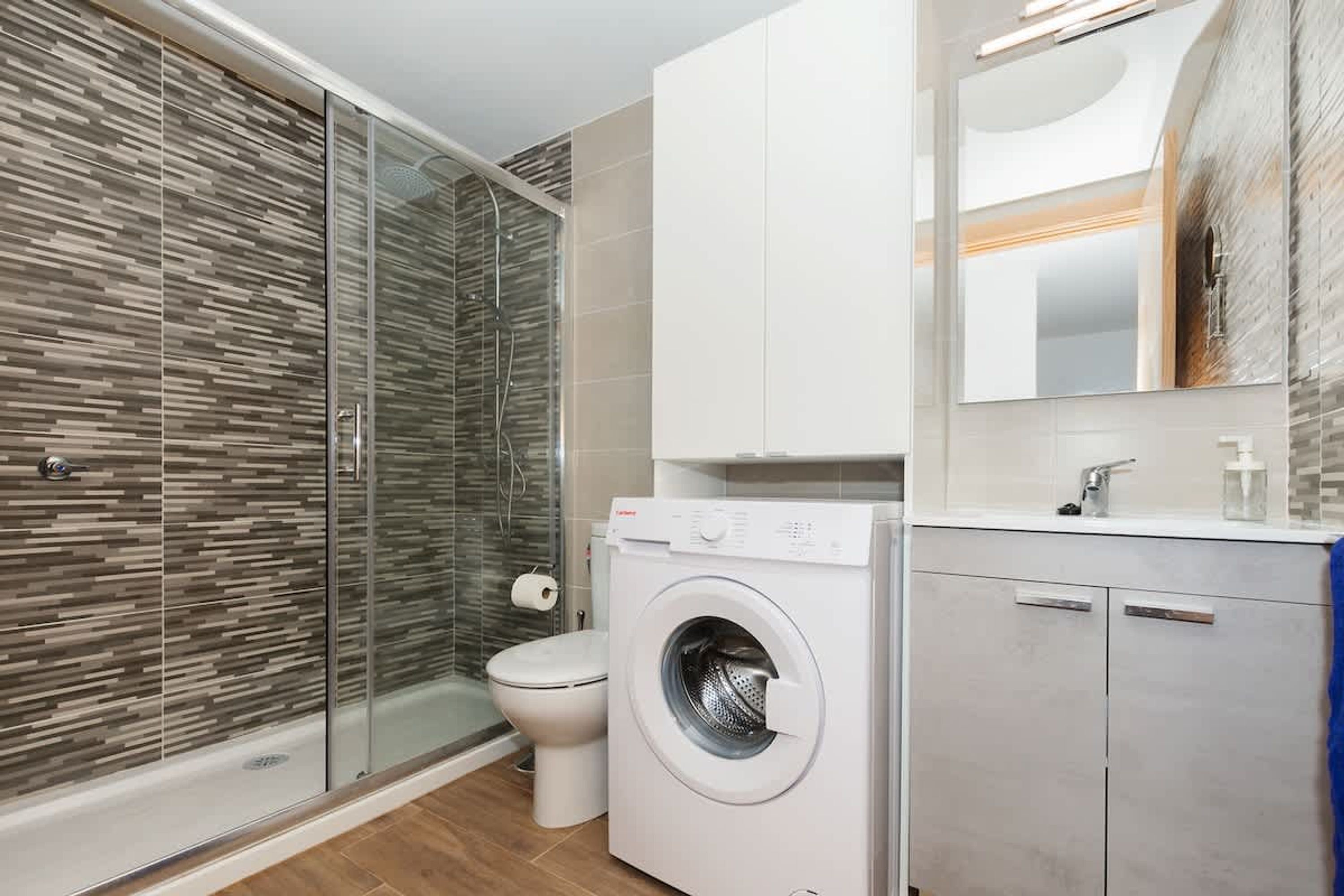 A modern bathroom with a glass shower enclosure, a built-in washing machine, and a large mirror above the sink.