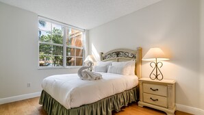 Un dormitorio con una cama grande, una mesita de noche con lámpara, una ventana con vista a árboles y un techo blanco.