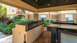 Un área de cocina al aire libre con una parrilla empotrada, rodeada de follaje y un edificio al fondo.