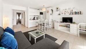 Een moderne woonkamer met een bank, salontafel en televisie. Er is een keuken met witte kasten en een eettafel met stoelen.