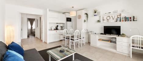 Un salon moderne avec un canapé, une table basse et un téléviseur. Il y a également un coin cuisine avec des meubles blancs et une table à manger avec des chaises.