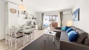 Een moderne woonkamer met een glazen eettafel, witte stoelen, een grijze bank met blauwe kussens, en een groot raam met uitzicht op het omliggende landschap.