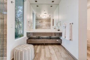 Une salle de bain moderne avec un grand miroir, deux lavabos, un banc et une vue sur les arbres par une fenêtre.