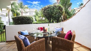 Mesa redonda de vidrio con sillas de mimbre, una botella y vasos en un patio con vistas a edificios y vegetación.