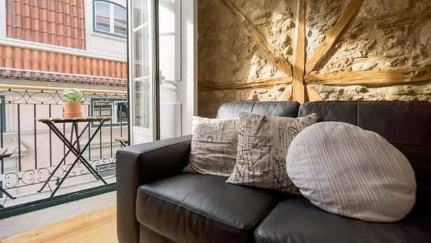 A cozy living room with a black leather sofa, a small wooden table with a potted plant, and a balcony with a metal railing. - Old Town Chiado Balcony Aircon by LD Apartments (Lisbon)