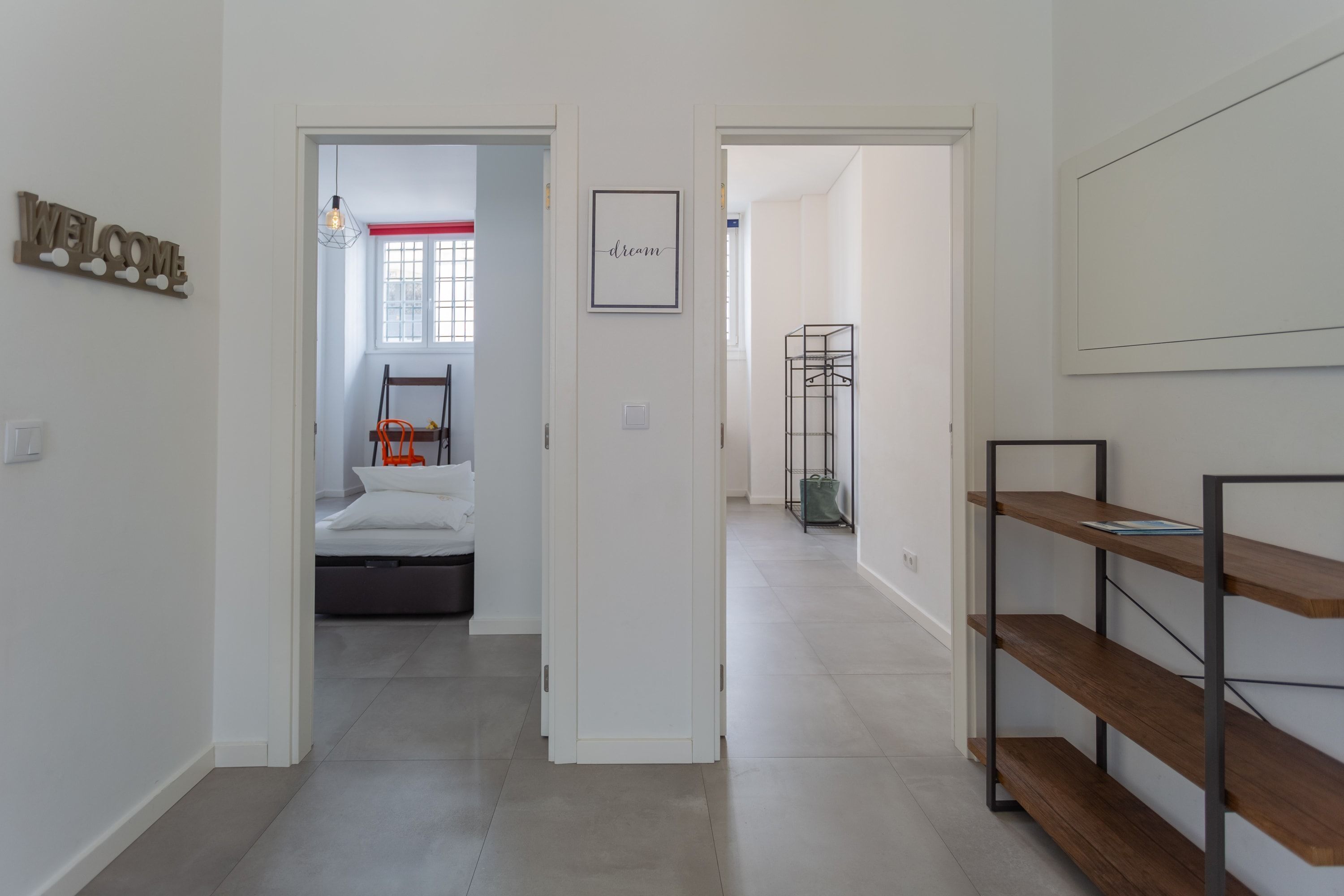 A hallway with a 'WELCOME' sign, a framed 'dream' sign, a bed with a red-striped headboard, and a metal shelving unit.