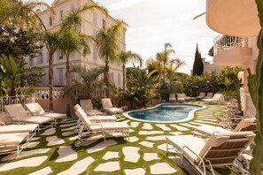 Un espace aménagé au bord de la piscine, avec des chaises longues et une piscine, le tout entouré de palmiers et d’un bâtiment à l’architecture classique.