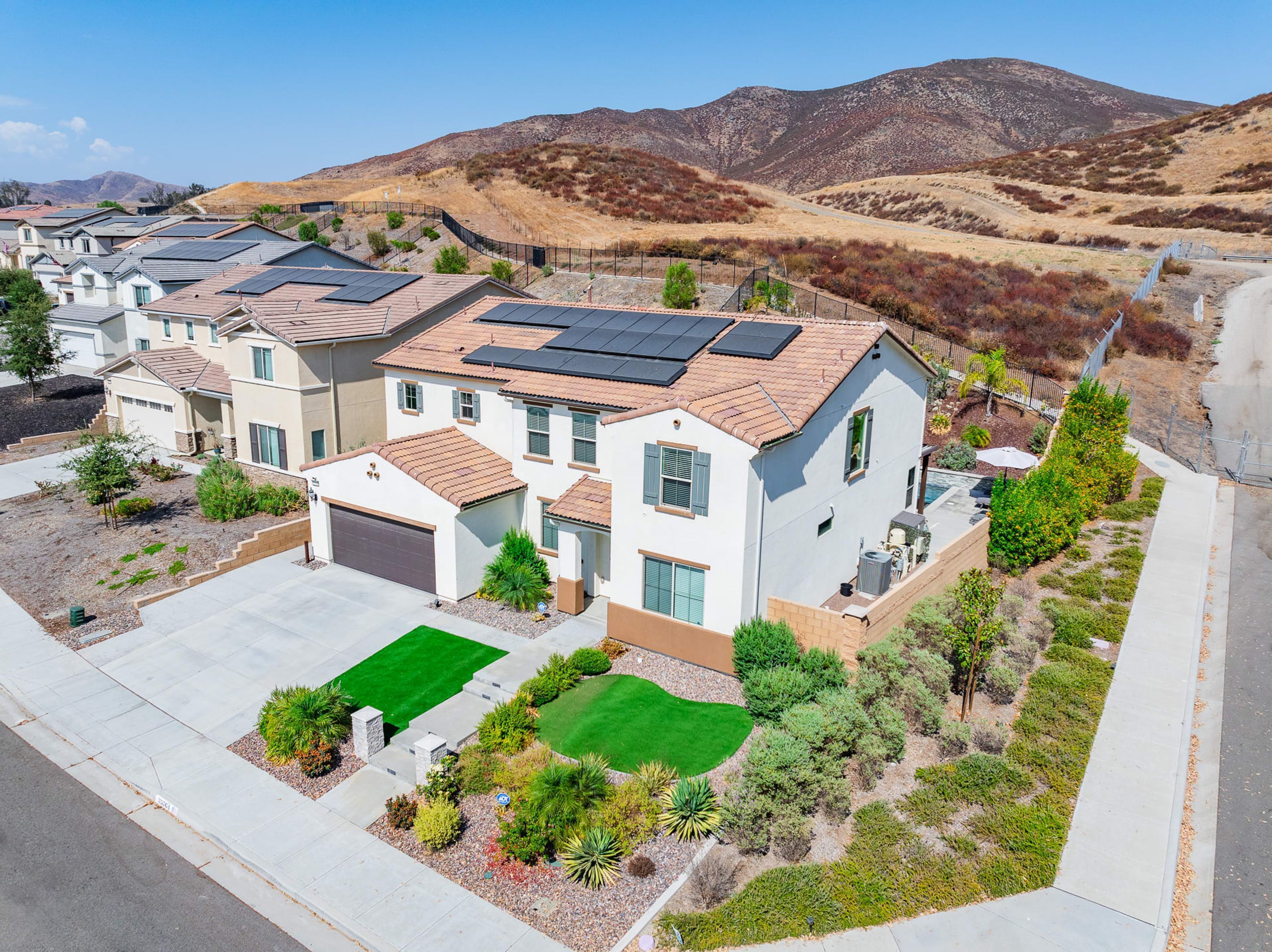 A residential area with houses featuring solar panels, green lawns, and a hilly backdrop.