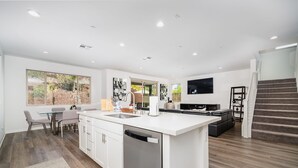 A modern kitchen with white cabinetry, a central island, and a built-in dishwasher. There is a dining area with a table and chairs, and a large window with a view of greenery. - Allura Champagne New in Wine Country Pool Game Rm (Winchester)