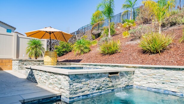 A swimming pool with stone walls and a waterfall feature, surrounded by landscaped plants and a patio area with an umbrella. - Allura Champagne New in Wine Country Pool Game Rm (Winchester)