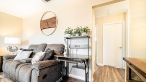 A living room with a gray sofa, a wooden coffee table, a wall clock, and a small shelf with plants. - Lux - Charming San Marcos 2BR Patio Pet-friendly (San Marcos)